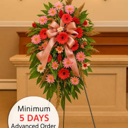 Tall sympathy spray of red and pink daisies with a peach bow on a metal easel in a chapel.