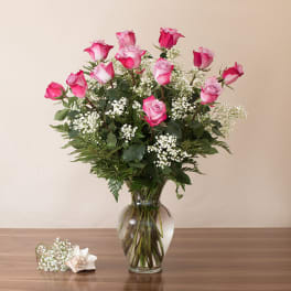Tall arrangement of pink roses with white filler flowers in a clear glass vase on a wooden table