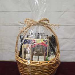 Large wicker gift basket wrapped in clear cellophane and tied with a raffia bow, filled with assorted gourmet items.