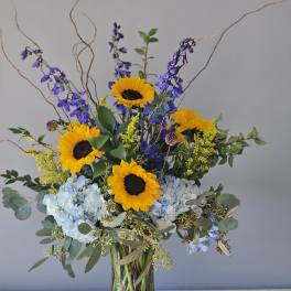 Tall arrangement of yellow sunflowers, blue hydrangeas, and delphinium in a clear glass vase