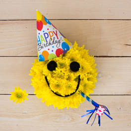 Round yellow mum arrangement shaped like a smiley face with birthday hat and party blower