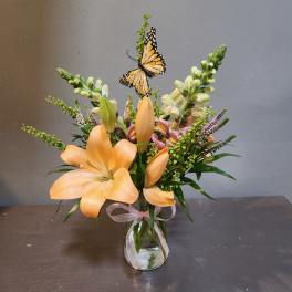 Peach lilies and mixed blooms in a glass vase with a decorative butterfly accent
