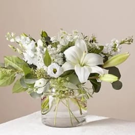 White lilies and mixed white flowers with lush greenery in a clear glass vase.