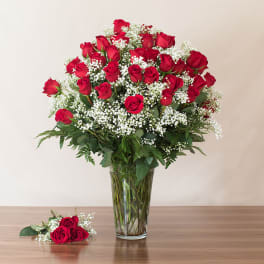 Tall arrangement of red roses and white filler flowers in a clear glass vase with a small rose bundle beside it.