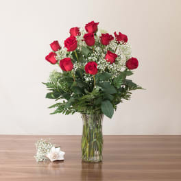 Tall arrangement of red roses with white filler flowers in a clear glass vase on a table.