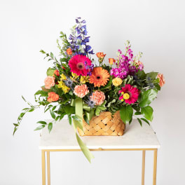 Colorful mixed flower basket with pink gerberas, orange carnations, and blue delphinium stems.