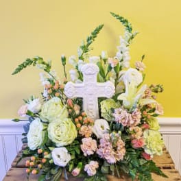 Pastel floral arrangement with white cross, pale green roses, lilies, and peach flowers.