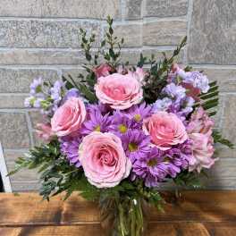 Pink roses and purple daisies arranged in a clear glass vase on a wooden surface.