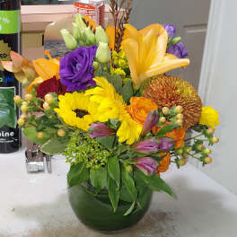 Bright mixed bouquet of yellow, orange, and purple flowers in a round green glass vase