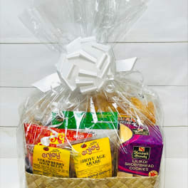 Woven gift basket wrapped in clear cellophane, filled with assorted colorful snack packages and topped with a white bow.