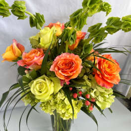 Tall arrangement of orange and green roses, green hydrangeas, and bells of Ireland in a clear glass vase