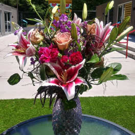 Tall arrangement of pink lilies, peach roses, and bright carnations in a purple textured glass vase