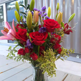 Tall bouquet of red roses, pink lilies, and purple accent flowers in a clear glass vase.