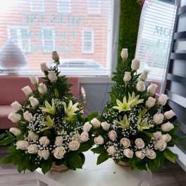Pair of white rose and lily arrangements with baby's breath in woven baskets on a white table.