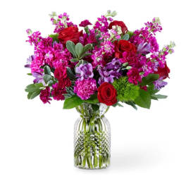 Jewel-tone bouquet of red roses, pink carnations, and purple blooms in a clear glass vase.