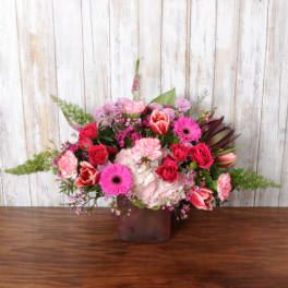Bright pink and red mixed flower arrangement in a frosted cube vase on a wooden table.