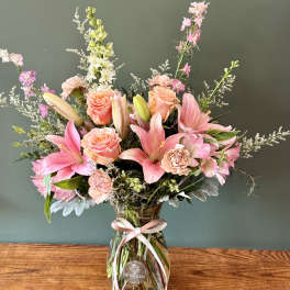 Tall bouquet of pink lilies, peach roses, and mixed blooms in a clear glass vase with a pink ribbon.