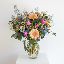 Mixed bouquet of peach gerberas, purple tulips, and blue flowers in a clear glass vase.
