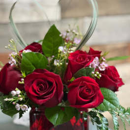 Short arrangement of red roses with small pink blossoms in a red glass cube vase with a heart-shaped grass accent.