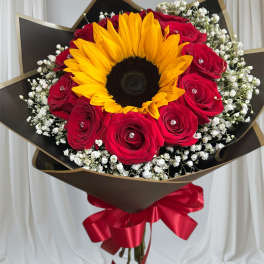 Bouquet with a large sunflower, red roses, and white filler flowers wrapped in dark paper with a red ribbon.