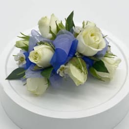 Small cluster of white roses with blue ribbon arranged as a corsage on a white stand
