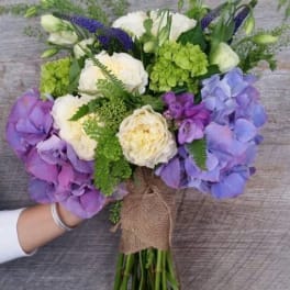 Hand-tied bouquet of cream blooms with purple hydrangeas and green accents wrapped in burlap