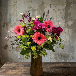 Tall bouquet of pink gerbera daisies and mixed blooms in an amber glass vase on a rustic wooden table.