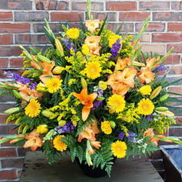 Tall arrangement of yellow gerbera daisies, orange lilies, and mixed flowers in a black urn.