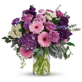 Mixed bouquet of pink gerbera daisies, lavender roses, and purple blooms in a clear glass vase