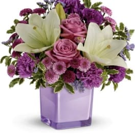 Lavender and purple arrangement with lilies, roses, carnations, and mums in a square lavender glass vase
