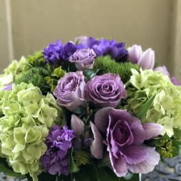 Low arrangement of lavender roses, green hydrangeas, and purple blooms in a white bowl