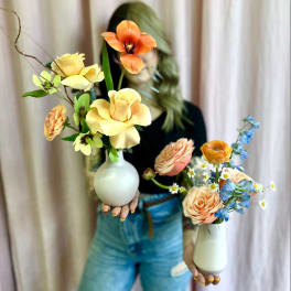 Two small bud vases with peach, yellow, orange, and blue flowers held by a person