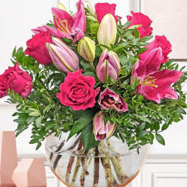 Arrangement of hot pink roses and pink lilies in a round clear glass vase on a wooden shelf