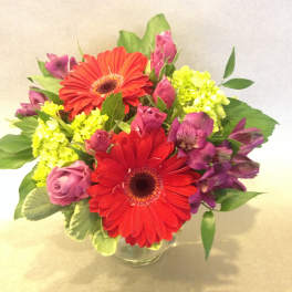 Bouquet of red gerbera daisies, pink roses, and purple flowers in a glass vase