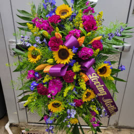 Standing spray of sunflowers, lilies, and purple flowers with a mourning ribbon
