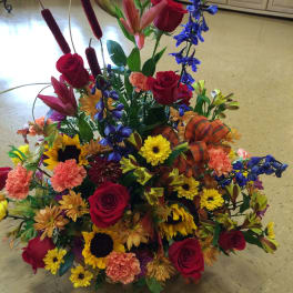 Tall mixed arrangement of red roses, sunflowers, lilies, and fall-colored blooms with a plaid bow.