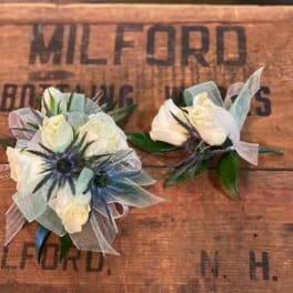 Two white floral boutonnieres with blue accents on a wooden surface