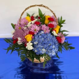 Basket arrangement of blue hydrangeas, pink daisies, and yellow roses
