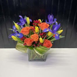 Bright arrangement of orange roses, purple iris, yellow tulips, and red berries in a clear glass cube vase