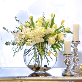 White and yellow floral arrangement in a round glass vase beside candles