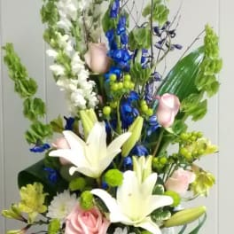 Tall floral arrangement with white lilies, pink roses, and blue flowers in a wooden vase