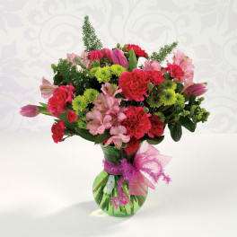 Bright pink carnations, alstroemeria, and green mums in a green glass vase with a pink bow