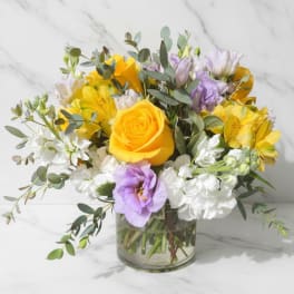 Low glass vase of yellow roses, lavender lisianthus, white stock, and yellow alstroemeria