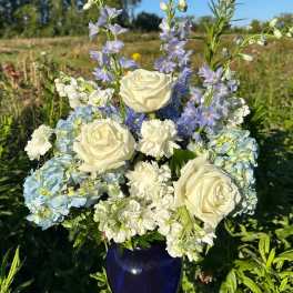 White roses, carnations, and blue hydrangeas with blue delphinium in a dark blue vase outdoors.