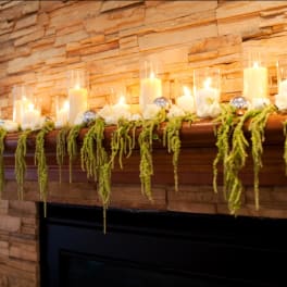 Candles and white roses arranged on a fireplace mantel with hanging greenery