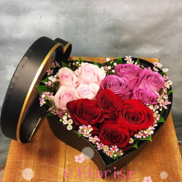 Heart-shaped black box filled with red, pink, and lavender roses and tiny pink filler flowers.