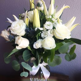 Tall arrangement of white lilies and roses with small white blooms in a clear glass vase with a white bow