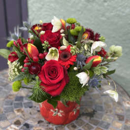 Compact arrangement of red roses, tulips, and mixed blooms in a red ceramic vase on a mosaic table.