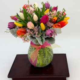 Mixed-color tulip arrangement with pink filler flowers in a green textured vase tied with a ribbon.