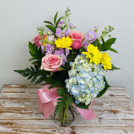 Mixed bouquet of pink roses, yellow daisies, blue hydrangea and lavender stems in a clear vase with pink ribbon
